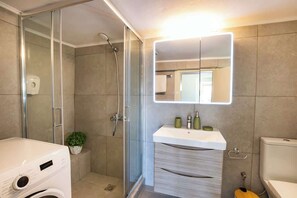 Une salle de bain moderne dotée d’une douche à parois de verre, d’un lave-linge encastré, d’un meuble-lavabo avec miroir et de toilettes.