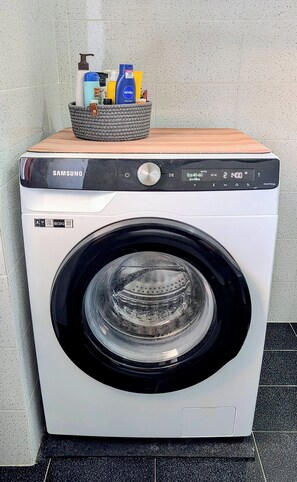 Una lavadora Samsung con panel de control, una cesta de detergente para ropa y un estante de madera encima.