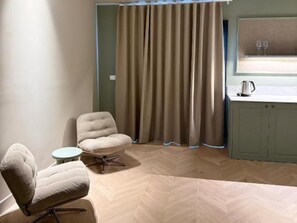 Ein Zimmer mit einem gepolsterten Sessel, einem kleinen runden Tisch, einem Fischgrätparkett, einem Vorhang und einer Küchenzeile mit Wasserkocher.