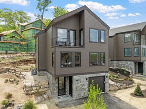 A modern multi-story house with a balcony, stone foundation, and a landscaped yard.