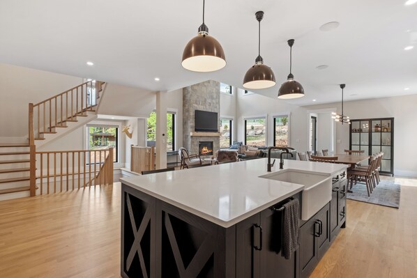 A spacious kitchen with a large island, wooden staircase, and a stone fireplace.