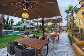 Un patio al aire libre con mesas, sillas, sombrillas y una piscina.