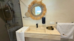 A bathroom with a glass shower enclosure, a marble sink, a round mirror with a straw-like frame, a black soap dispenser, and a towel rack with a white towel. - Jnane Rihane Marrakech (Marrakech)
