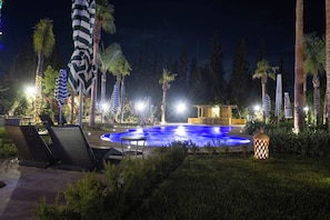 Ein Poolbereich bei Nacht mit Liegestühlen, Sonnenschirmen und einer Fontäne.