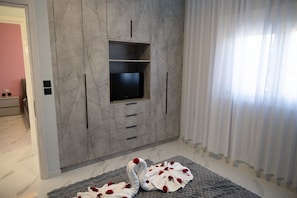 A modern living room with a flat-screen TV mounted on a marble wall, a grey rug with red flowers, and a window with sheer curtains. - Merdanai Villa (64C3+69, Archaggelos 851 02, Greece)