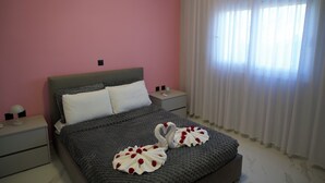 Ein Schlafzimmer mit Bett, Kissen, einem Nachttisch, einer Lampe und einem Fenster mit Vorhängen.