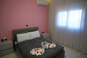 A bedroom with a bed, pillows, a nightstand, a lamp, and a window with curtains. - Merdanai Villa (64C3+69, Archaggelos 851 02, Greece)
