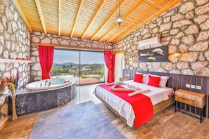 A bedroom with a stone wall, a wooden ceiling, a bed with red bedding, a bathtub, and a view of the landscape.