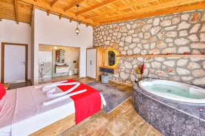 A room with a stone wall, a wooden ceiling, a large bathtub, and a bed with a red blanket.