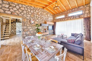 A stone-walled room with a dining table set for four, a sofa, a TV, and a staircase.