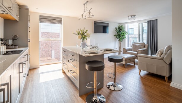 A modern kitchen with a central island, bar stools, a sofa, and a TV mounted on the wall. - Swift Apartments - Apartment 6 with Free Gym Access (York)