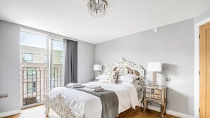 A bedroom with a large bed, a nightstand, a chandelier, and a view of a building through a window. - Swift Apartments - Apartment 6 with Free Gym Access (York)