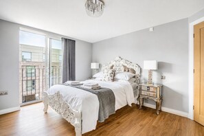 Ein Schlafzimmer mit einem großen Bett, einem Nachttisch, einer Pendelleuchte und Blick auf ein Gebäude durch ein Fenster.