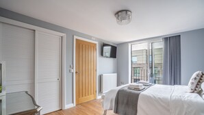 A bedroom with a bed, a television, a wooden door, and a window with a view. - Swift Apartments - Apartment 6 with Free Gym Access (York)