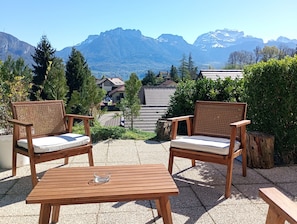 Dos sillas de mimbre con cojines, una mesa de madera y una vista de montañas y casas al fondo.