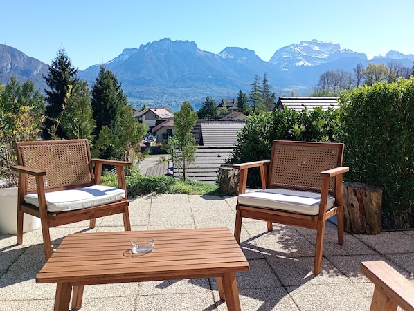 Two wicker chairs with cushions, a wooden table, and a view of mountains and houses in the background.