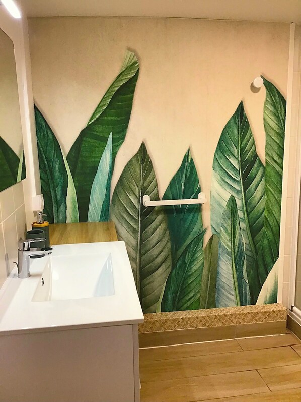 Un baño con un espejo grande, un lavabo blanco y un mural de hojas verdes.