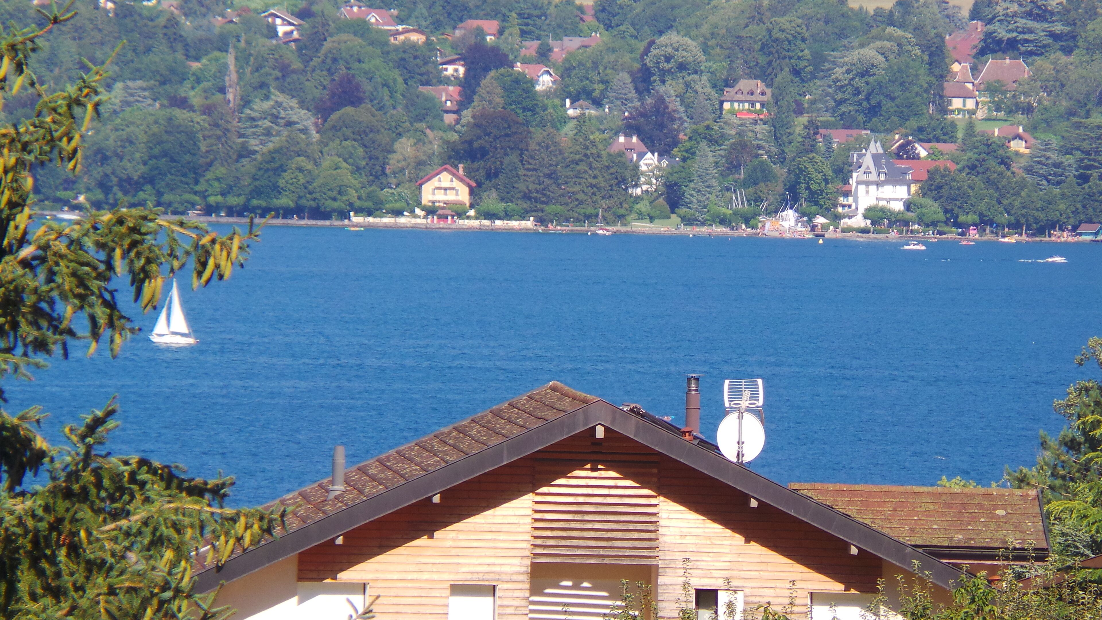 A lakeside view with a wooden house in the foreground, a sailboat on the water, and houses in the background.