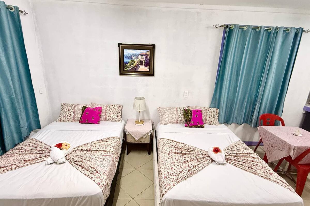 A room with two beds, a small table, a chair, a lamp, and a framed picture on the wall.