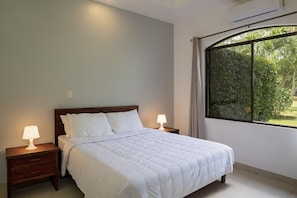 A bedroom with a bed, two bedside lamps, a window with a view of greenery, and an air conditioning unit.
