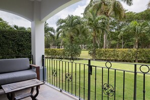A balcony with a grey sofa and a wooden table, overlooking a well-maintained garden with palm trees and hedges.