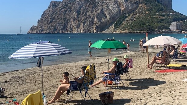 Ein Strand mit Menschen, die unter Sonnenschirmen entspannen, ein felsiger Berg im Hintergrund und ein klarer, blauer Himmel.