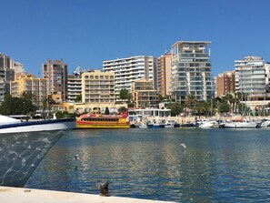 Ein Yachthafen mit Booten, einer gelb-roten Fähre und modernen Gebäuden im Hintergrund.