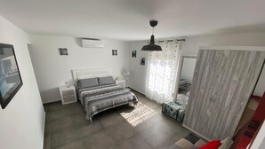 Ein Schlafzimmer mit einem Bett, Nachttischen, einem Ventilator, einem Fenster mit Vorhängen und einem roten Kissen auf einem Sofa.