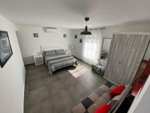 Ein Schlafzimmer mit einem Bett, Nachttischen, einem Ventilator, einem Fenster mit Vorhängen und einem roten Kissen auf einem Sofa.