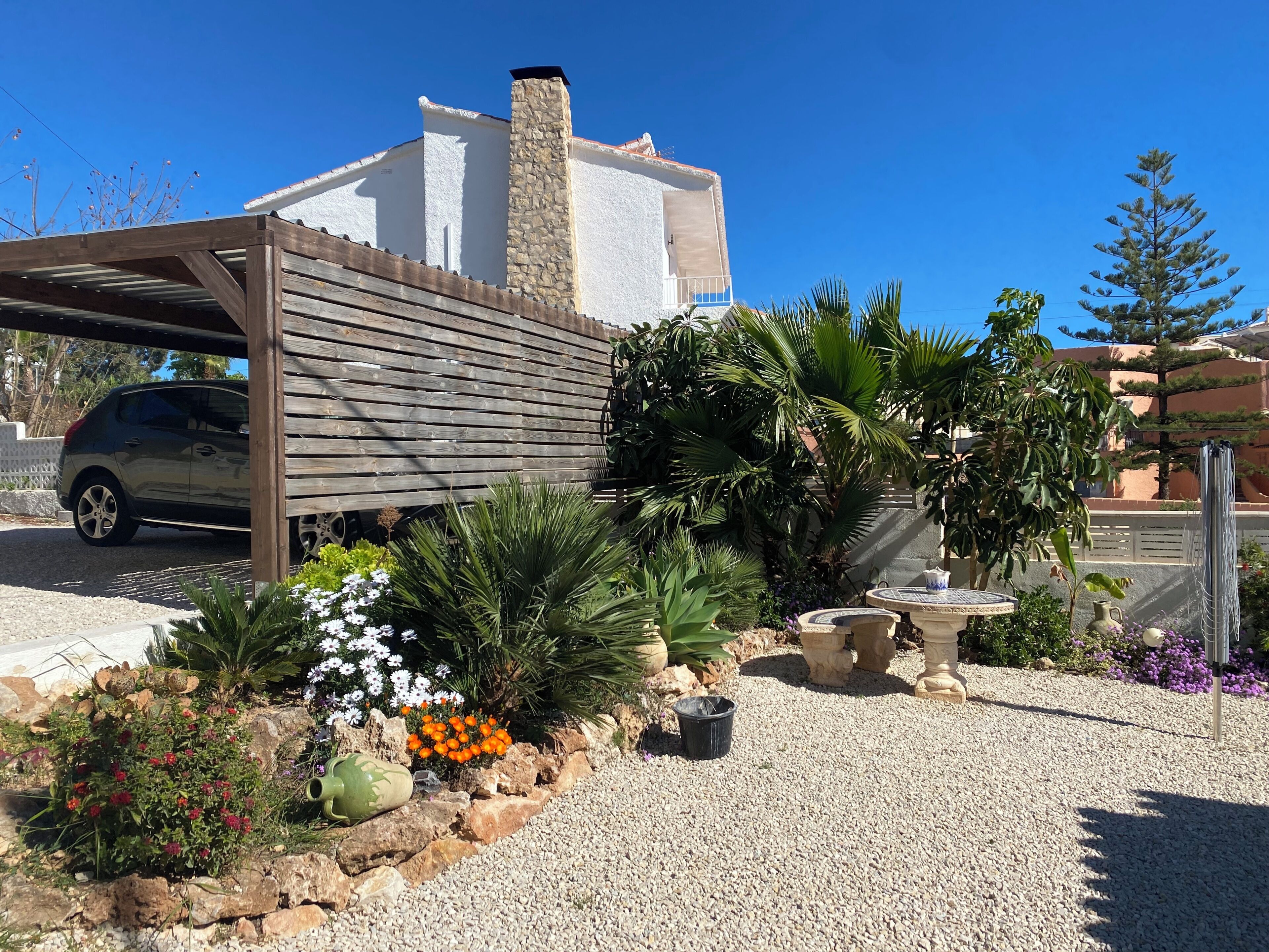 A house with a covered carport, a stone chimney, and a garden with various plants and flowers.