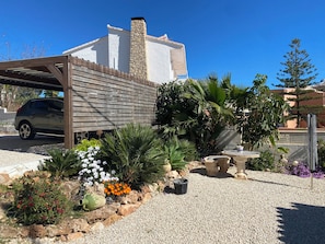 Ein Haus mit überdachter Auffahrt, einem Steinsschornstein und einem Garten mit verschiedenen Pflanzen und Blumen.