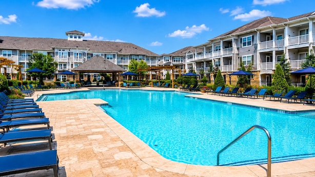 A swimming pool with lounge chairs and umbrellas, surrounded by multi-story buildings with balconies. - Elite Cozy Suites Buford (Buford)