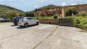 A parking area with a white car and a boat trailer, a building with a staircase, and a hillside in the background. - Terrace With sea View, Parking Space, air Conditioning (Castelsardo)