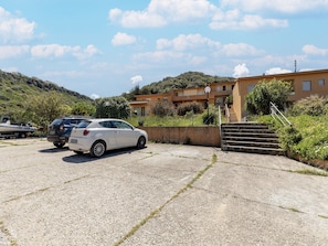 A parking area with a white car and a boat trailer, a building with a staircase, and a hillside in the background. - Terrace With sea View, Parking Space, air Conditioning (Castelsardo)