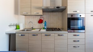A modern kitchen with a gas stove, oven, and stainless steel appliances. There are wooden cabinets with handles, a tiled backsplash, and a hanging oven mitt. - Terrace With sea View, Parking Space, air Conditioning (Castelsardo)