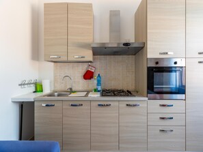 A modern kitchen with a gas stove, oven, and stainless steel appliances. There are wooden cabinets with handles, a tiled backsplash, and a hanging oven mitt. - Terrace With sea View, Parking Space, air Conditioning (Castelsardo)