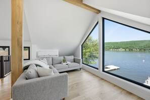 Ett modernt vardagsrum med ett stort fönster med utsikt över en sjö, en grå soffa och en synlig takbalk i trä.