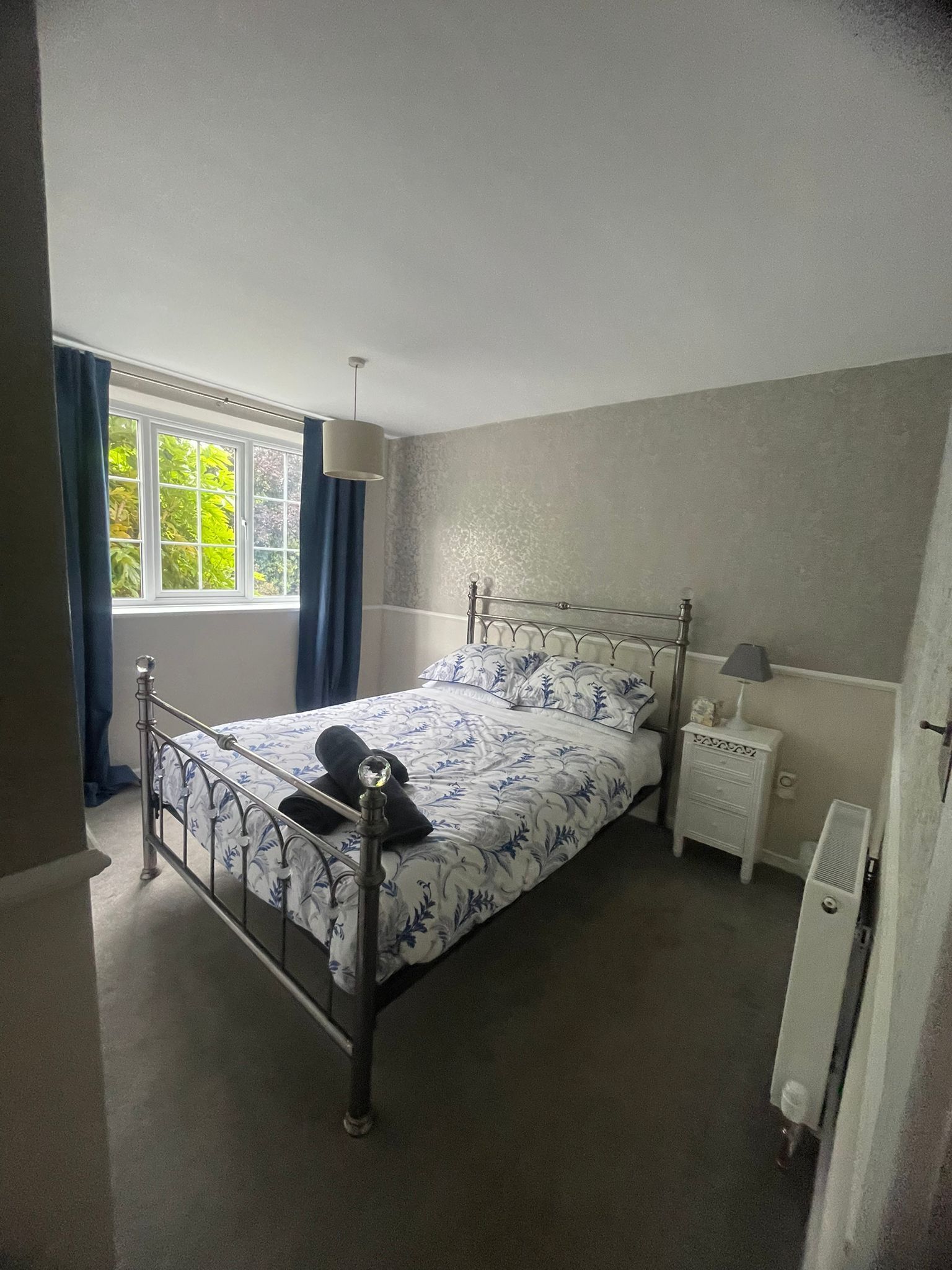 A bedroom with a metal bed frame, a bedside table with a lamp, a window with curtains, and a radiator.