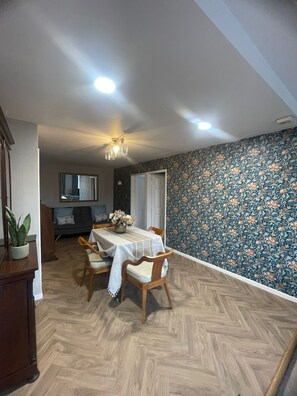 A dining area with a table set for four, wooden chairs, a chandelier, and a patterned wallpaper wall. - Carnegie Library Christie apartment (Swinton)