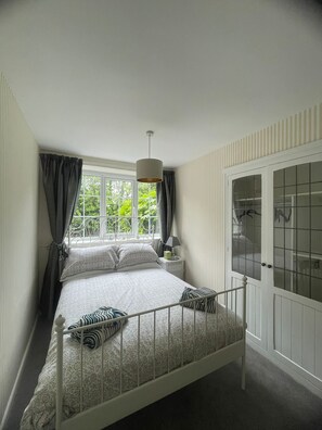 A bedroom with a bed, a nightstand, a lamp, a window with curtains, and a wardrobe. - Carnegie Library Christie apartment (Swinton)