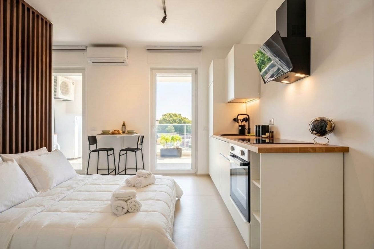 A modern hotel room with a bed, a small kitchenette, a dining area, and a view of the outdoors.