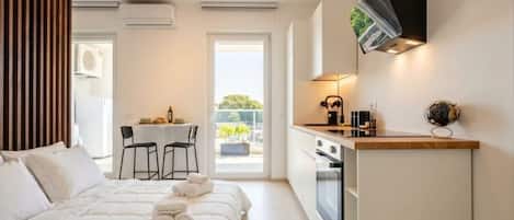 A modern hotel room with a bed, a small kitchenette, a dining area, and a view of the outdoors.