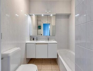 A bathroom with white tiles, a sink, a mirror, and a bathtub. - Sea View Apartment for 6 in Zeedijk (Blankenberge)