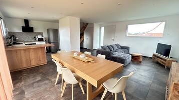A modern kitchen with a wooden island, a dining area with a wooden table and chairs, a grey sectional sofa, and a TV on a wooden stand.