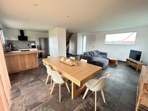 A modern kitchen with a wooden island, a dining area with a wooden table and chairs, a grey sectional sofa, and a TV on a wooden stand.