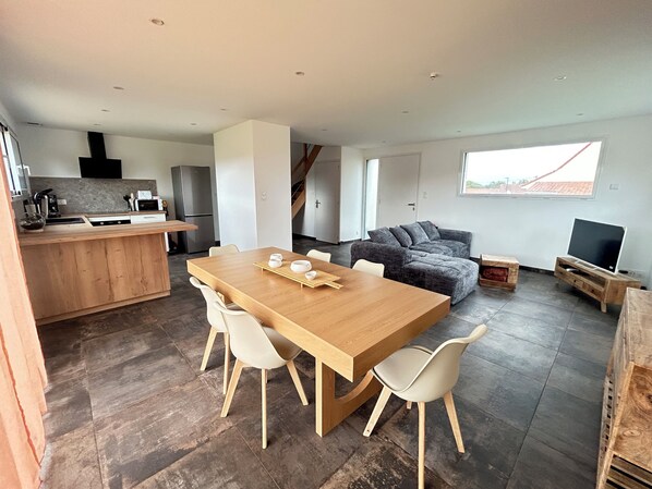 Una cocina moderna con una isla de madera, un comedor con mesa y sillas de madera, un sofá seccional gris y un televisor sobre un mueble de madera.