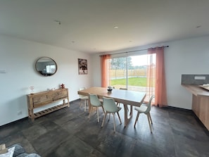 A dining area with a wooden table and chairs, a wooden cabinet, a round mirror, and a view of a backyard through large windows.