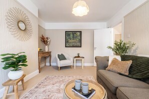 Una sala de estar con un sofá, una mesa de centro con tazas y un libro, una silla, una mesita con un florero y un cuadro en la pared.