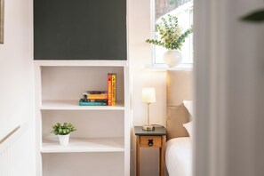 Ein Schlafzimmer mit Nachttisch, Lampe, einem Fenster mit Pflanze und einem Bücherregal mit Büchern.
