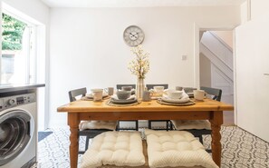 A dining area with a wooden table set for four, a clock on the wall, and a washing machine in the background.